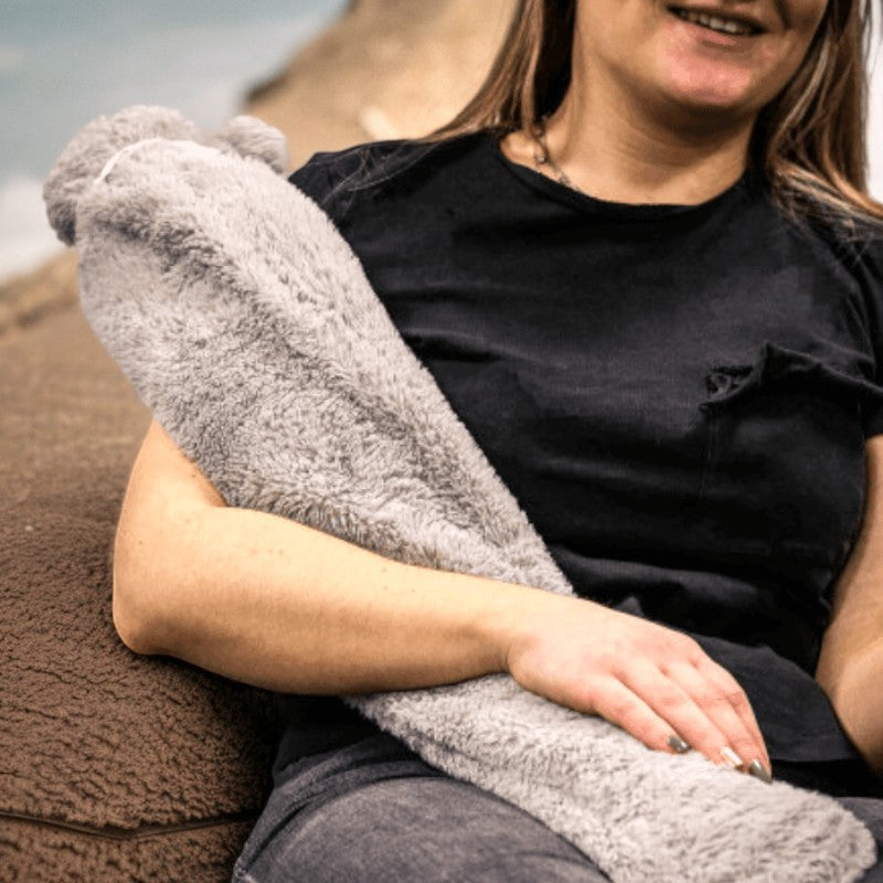 Long hot water bottle in grey fleece cover resting on a person's lap