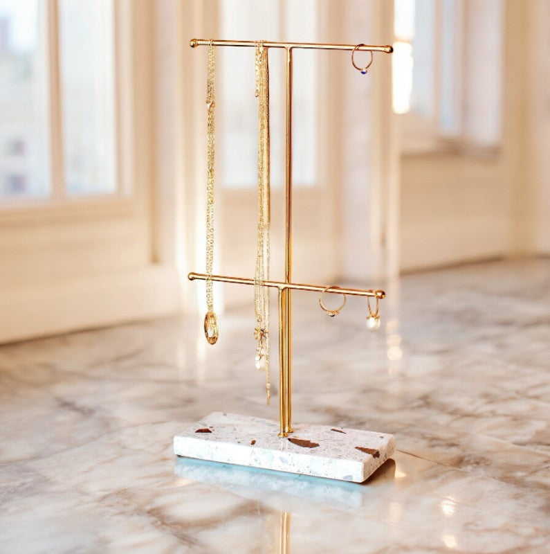 Gold two-tier jewellery stand with a terrazzo-style base displaying jewellery by a window