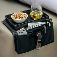 Black sofa armrest organiser with top tray and side pockets holding remotes and reading material