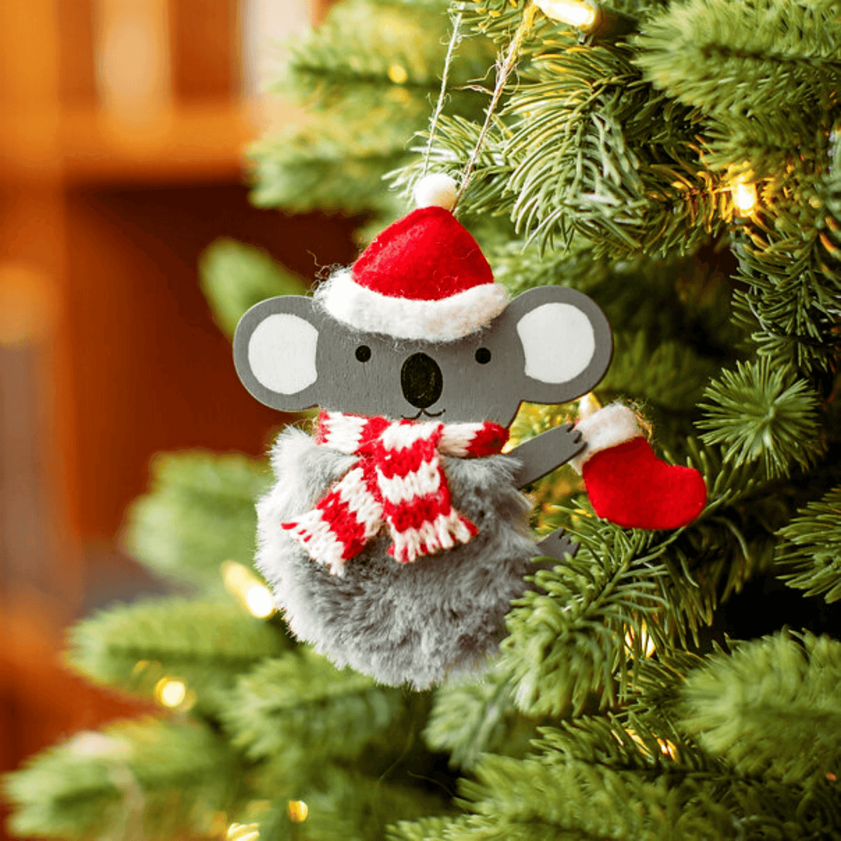 Festive koala ornament with fluffy grey body, Santa hat and striped scarf hanging on a Christmas tree