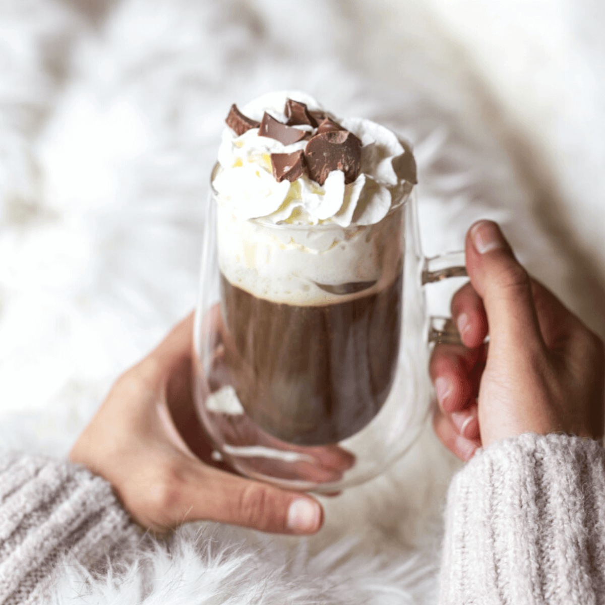 Hands holding a double-walled glass mug topped with whipped cream